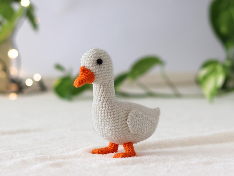 Puede incluir: Un ganso blanco de crochet con pico y patas naranjas. El ganso est&aacute; de pie sobre una superficie blanca.
