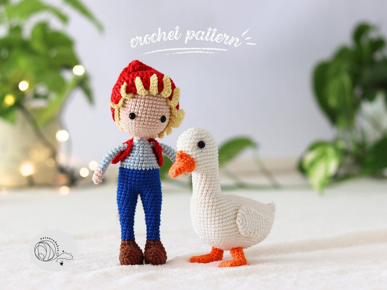 Puede incluir: Un mu&ntilde;eco de crochet de ni&ntilde;o con un sombrero rojo y un mono azul est&aacute; de pie junto a un mu&ntilde;eco de ganso de crochet blanco. El mu&ntilde;eco de ni&ntilde;o tiene el pelo rubio y los ojos marrones. El mu&ntilde;eco de ganso tiene un pico y patas naranjas.