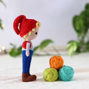 Puede incluir: Un mu&ntilde;eco de gnomo de crochet con un gorro rojo, pantalones azules y botas marrones se encuentra junto a tres ovillos de lana en naranja, verde y azul.