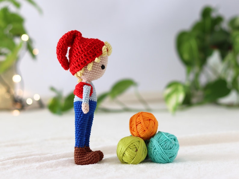 Puede incluir: Un mu&ntilde;eco de gnomo de crochet con un gorro rojo, pantalones azules y botas marrones se encuentra junto a tres ovillos de lana en naranja, verde y azul.