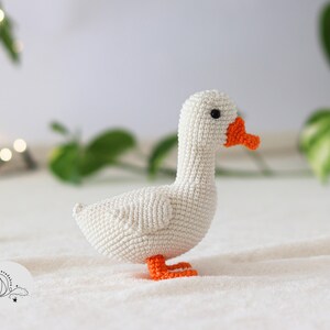 Puede incluir: Un ganso blanco de crochet con pico y patas naranjas. El ganso est&aacute; sentado sobre una superficie blanca y esponjosa.