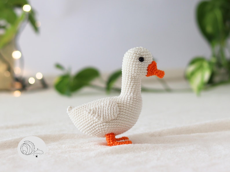 Puede incluir: Un ganso blanco de crochet con pico y patas naranjas. El ganso est&aacute; sentado sobre una superficie blanca y esponjosa.