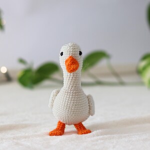 Puede incluir: Un juguete de ganso blanco de crochet con patas y pico naranja. El ganso est&aacute; de pie sobre una superficie blanca.