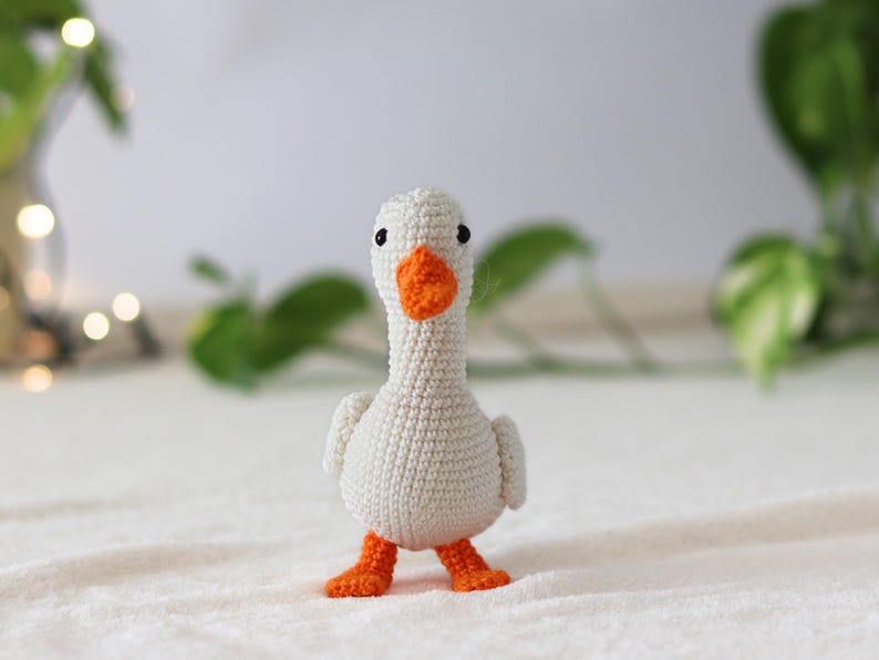 Puede incluir: Un juguete de ganso blanco de crochet con patas y pico naranja. El ganso est&aacute; de pie sobre una superficie blanca.