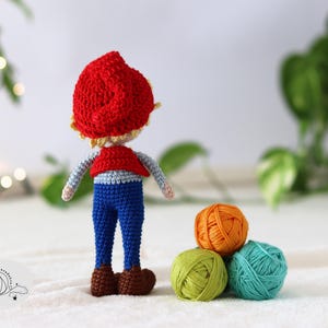 Puede incluir: Una mu&ntilde;eca de crochet con un sombrero rojo, pantalones azules y un chaleco rojo. La mu&ntilde;eca est&aacute; de pie sobre una superficie blanca con tres ovillos de lana delante de ella.