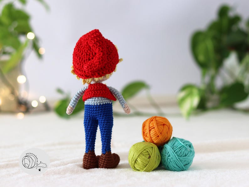 Puede incluir: Una mu&ntilde;eca de crochet con un sombrero rojo, pantalones azules y un chaleco rojo. La mu&ntilde;eca est&aacute; de pie sobre una superficie blanca con tres ovillos de lana delante de ella.
