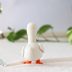 Puede incluir: Pato amigurumi blanco de crochet con patas naranjas, de pie sobre una superficie blanca.