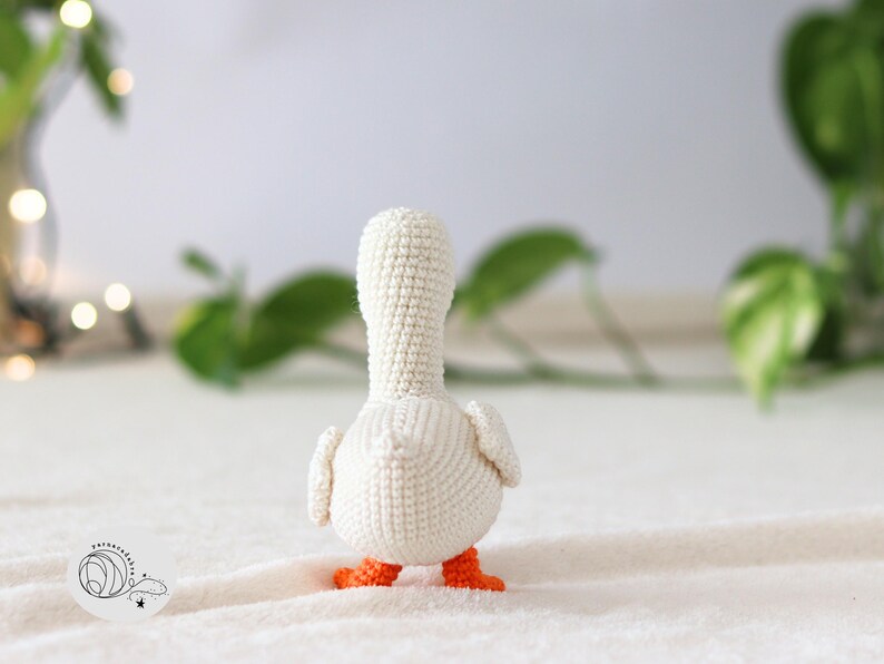 Puede incluir: Pato amigurumi blanco de crochet con patas naranjas, de pie sobre una superficie blanca.