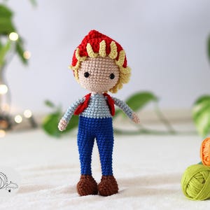 Puede incluir: Una mu&ntilde;eca de crochet con pelo rojo y amarillo, que lleva un chaleco rojo, pantalones vaqueros azules y zapatos marrones.
