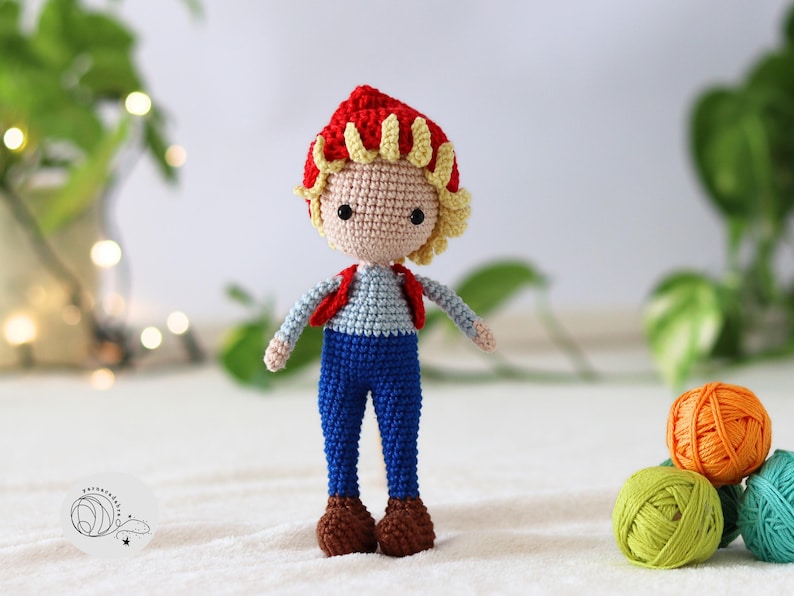 Puede incluir: Una mu&ntilde;eca de crochet con pelo rojo y amarillo, que lleva un chaleco rojo, pantalones vaqueros azules y zapatos marrones.