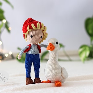 Puede incluir: Una mu&ntilde;eca de crochet con un sombrero rojo y un mono azul se encuentra junto a un ganso blanco de crochet con un pico naranja.