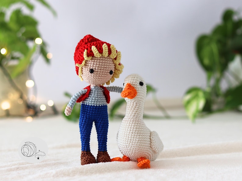 Puede incluir: Una mu&ntilde;eca de crochet con un sombrero rojo y un mono azul se encuentra junto a un ganso blanco de crochet con un pico naranja.
