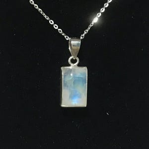 May include: A silver necklace with a rectangular moonstone pendant. The moonstone has a blue and white iridescent glow. The chain is delicate and the pendant is set in a silver bezel. The background is black.