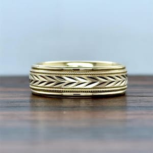 May include: A gold band ring featuring a central horizontal band with a repeating chevron or leaf pattern. The central design is bordered on the top and bottom by thin, raised beaded edges, creating a textured and detailed metallic finish.