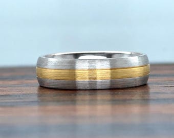 May include: A two-tone ring featuring a wide band with a brushed silver-colored finish on the outer edges and a central horizontal band with a brushed gold-colored finish. The ring has a flat profile and a consistent width throughout its circumference.