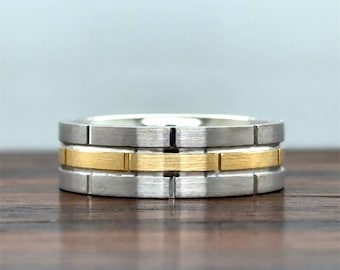 May include: A two-tone band ring featuring a brushed silver-colored exterior with a central horizontal band of brushed gold-colored metal. The ring has vertical grooves spaced evenly around the circumference, creating a segmented appearance.