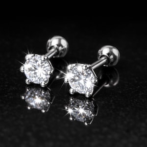 May include: A pair of silver-toned stud earrings featuring round, clear, faceted stones held in six-prong settings. Each earring has a smooth, spherical back closure attached to a straight post, reflecting light with a bright, sparkling effect.