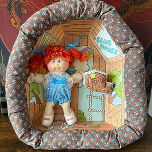 May include: A fabric-framed wall hanging featuring a doll with orange yarn hair and blue eyes, wearing a blue polka-dot romper with white lace trim. The background depicts a treehouse scene with text reading Team Club House.