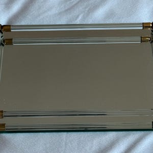 May include: Rectangular mirror with a reflective surface. The mirror is framed with clear, ribbed glass and gold-colored metal corner accents. The mirror is likely used for home decor.