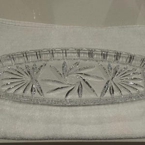 May include: A clear, rectangular crystal dish with a decorative starburst and leaf design. The dish is approximately 12 inches long and sits on a white towel. The edges of the dish have a ribbed pattern.