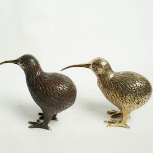 May include: Two bronze kiwi bird figurines, one with a brown finish and the other with a gold finish. The birds are standing on a white surface.