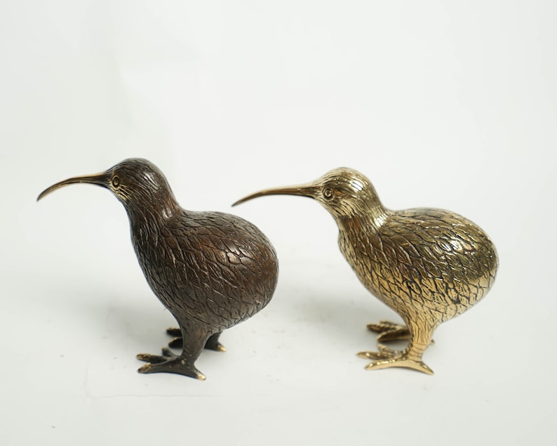 May include: Two bronze kiwi bird figurines, one with a brown finish and the other with a gold finish. The birds are standing on a white surface.