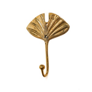 May include: A gold-toned metal hook shaped like a stylized ginkgo leaf. The hook is designed for hanging items on a wall.