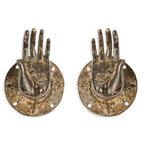 Może przedstawiać: Para antycznych mosiężnych klamki drzwiowych w kształcie dłoni z misternymi detalami. Klamki są zamontowane na okrągłych płytach z ozdobnymi krawędziami.