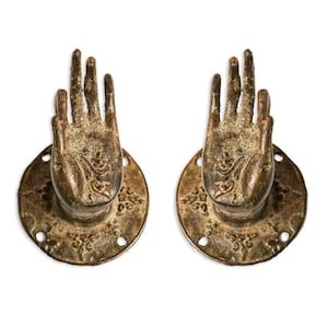 Może przedstawiać: Para antycznych mosiężnych klamki do drzwi w kształcie dłoni z misternymi wzorami kwiatowymi. Klamki są zamontowane na okrągłych płytach z ozdobnymi krawędziami.