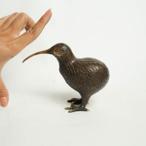 May include: A bronze sculpture of a kiwi bird. The bird is standing on a white surface and has a long, thin beak.