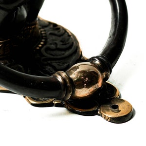 Puede incluir: Primer plano de un aldaba decorativa. La aldaba presenta un acabado en bronce oscuro con detalles dorados. El asa tiene forma de anillo curvado. La base tiene un dise&ntilde;o decorativo con un orificio para el montaje.