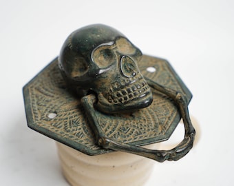 May include: A bronze skull door knocker with a detailed design. The skull is attached to a decorative octagonal base with a skeletal handle.