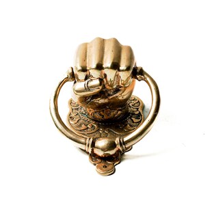 Puede incluir: Un aldaba de lat&oacute;n con forma de pu&ntilde;o cerrado. La aldaba tiene un asa de anillo y est&aacute; montada sobre una base decorativa con detalles ornamentados. El acabado es de color dorado brillante.