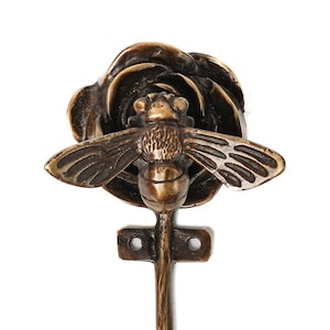 May include: A brass hook shaped like a rose with a bee perched on top. The hook is a decorative accent for a home or garden.