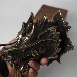 May include: A detailed brass dragon head sculpture with intricate details and a textured finish. The dragon's mouth is open, revealing sharp teeth, and its eyes are large and expressive.