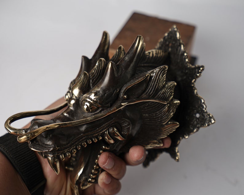May include: A detailed brass dragon head sculpture with intricate details and a textured finish. The dragon's mouth is open, revealing sharp teeth, and its eyes are large and expressive.