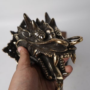 May include: A detailed brass dragon head door knocker with a textured surface and open mouth. The dragon has a gold ring on its nose and a decorative base.