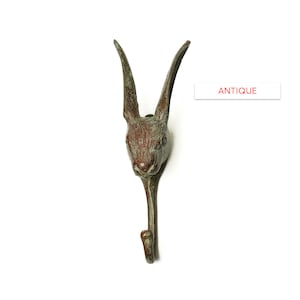 May include: Antique bronze rabbit head hook. The hook is shaped like a rabbit's head with long ears and a curved body. The hook is a dark brown color with a green patina.