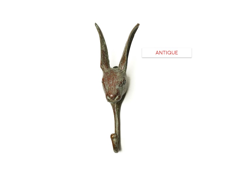 May include: Antique bronze rabbit head hook. The hook is shaped like a rabbit's head with long ears and a curved body. The hook is a dark brown color with a green patina.