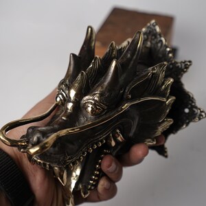 May include: A detailed brass dragon head door knocker with a gold finish. The dragon's mouth is open, revealing sharp teeth, and its eyes are inlaid with gold. The dragon's wings are spread out, and its tail is curled around its body.