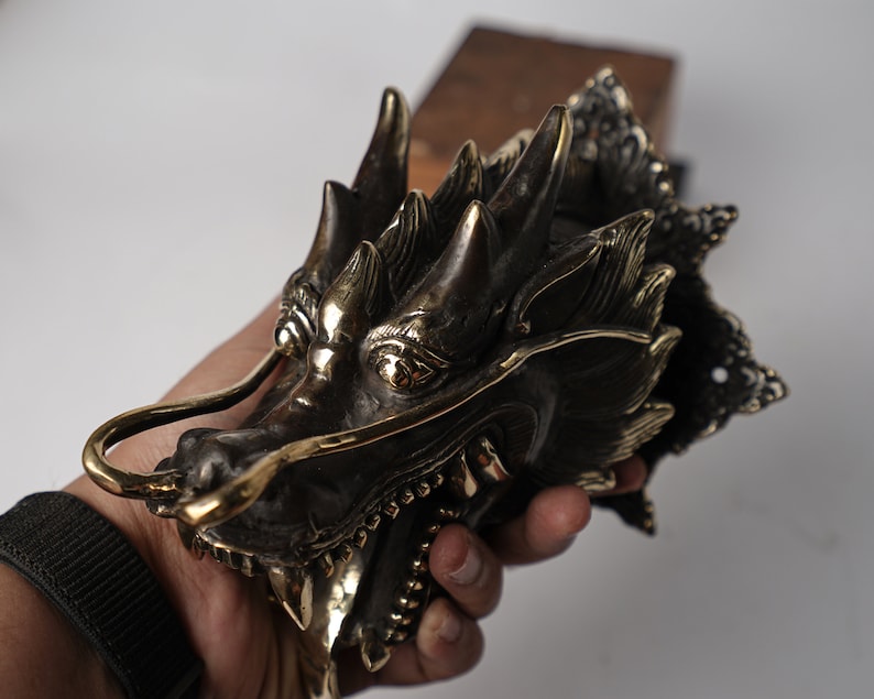 May include: A detailed brass dragon head door knocker with a gold finish. The dragon's mouth is open, revealing sharp teeth, and its eyes are inlaid with gold. The dragon's wings are spread out, and its tail is curled around its body.