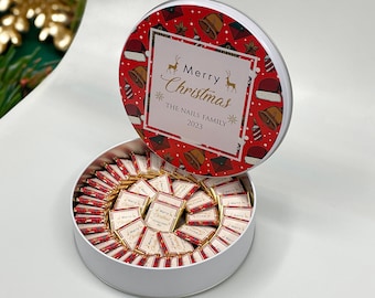 May include: A round white tin with a red lid. The lid has a Christmas design with Santa hats, bells, and reindeer. The text on the lid reads "Merry Christmas The Nails Family 2023". The tin is filled with small, individually wrapped chocolates.