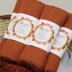 May include: Several rust-colored shawls with fringed edges are bundled together in a wicker basket. Each shawl is wrapped with a white label featuring the text "Thanks!" and a fall-themed leaf design. The text includes names and a date.