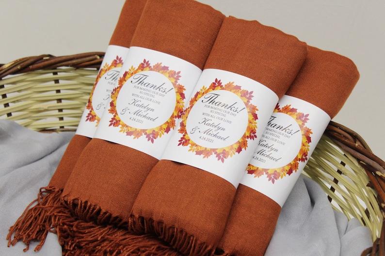 May include: Several rust-colored shawls with fringed edges are bundled together in a wicker basket. Each shawl is wrapped with a white label featuring the text "Thanks!" and a fall-themed leaf design. The text includes names and a date.
