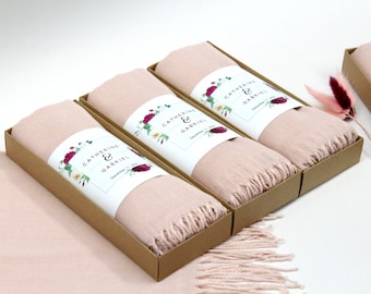 May include: Three pink scarves with fringed edges are packaged in brown boxes. Each box has a white label with a floral design and the text "Catherine & Gabriel, December 11, 2021".