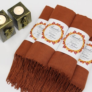 May include: Four rolled rust-colored scarves with fringe, each wrapped with a white label featuring the word "Thanks!" and a fall leaf design. Two gold-colored candle holders with lit candles are in the background.