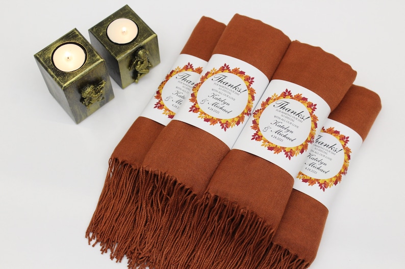May include: Four rolled rust-colored scarves with fringe, each wrapped with a white label featuring the word "Thanks!" and a fall leaf design. Two gold-colored candle holders with lit candles are in the background.