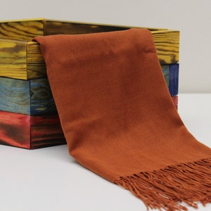 May include: A rust-colored scarf with fringe detail is draped over a stack of three wooden boxes. The boxes are painted in yellow, blue, and red hues. The scarf appears to be made of a soft fabric.