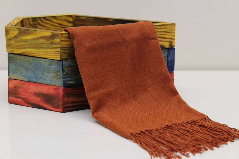 May include: A rust-colored scarf with fringe detail is draped over a stack of three wooden boxes. The boxes are painted in yellow, blue, and red hues. The scarf appears to be made of a soft fabric.