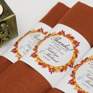 May include: Close-up of folded rust-colored napkins with white labels. The labels feature a fall leaf wreath design and the text "Thanks!" along with names and a date. A gold candle holder with an angel is in the background.